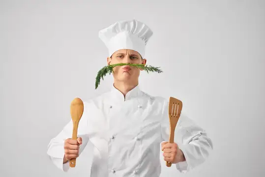Повар в колпаке с веточкой зелени в зубах и кухонными лопатками