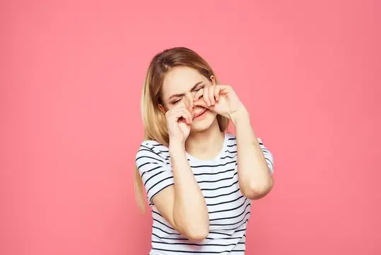 Светловолосая девушка в полосатой футболке трет глаза на розовом фоне