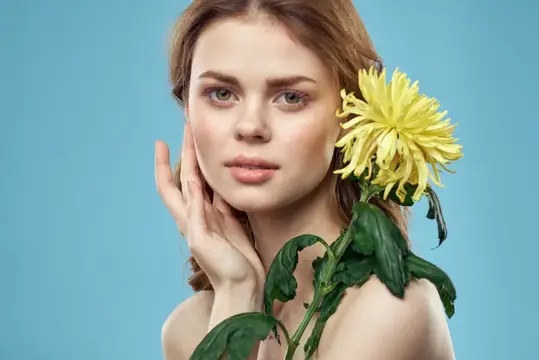 Красивая женщина с жёлтым цветком у лица, улыбающаяся, кадрированный портрет с обнажёнными плечами