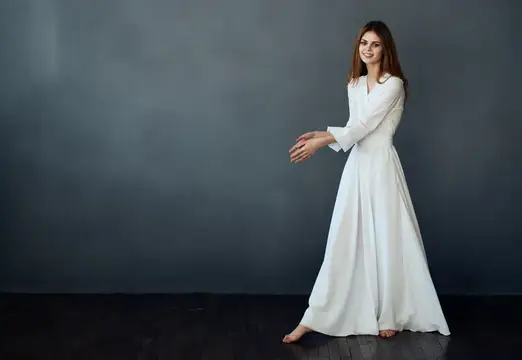 Улыбающаяся девушка в белом платье танцует у серой стены