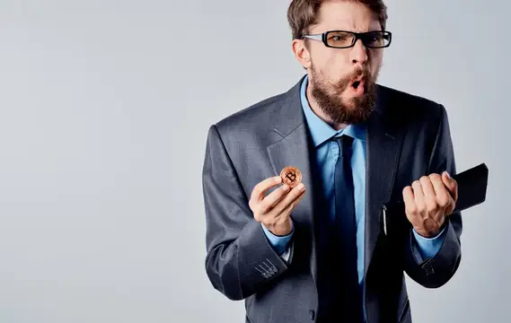 Бородатый мужчина в очках и костюме удивлённо смотрит на монету
