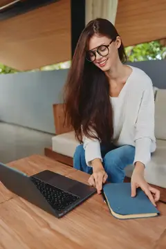 Улыбающаяся темноволосая девушка в очках работает за ноутбуком с блокнотом