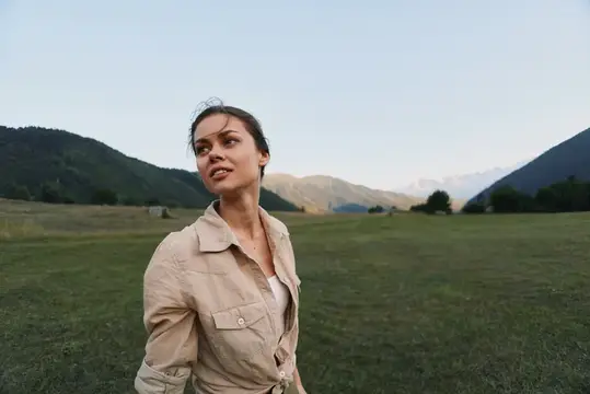 Молодая женщина в бежевой рубашке на фоне гор и луга