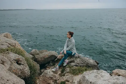 Девушка в сером свитере сидит на скалах у моря