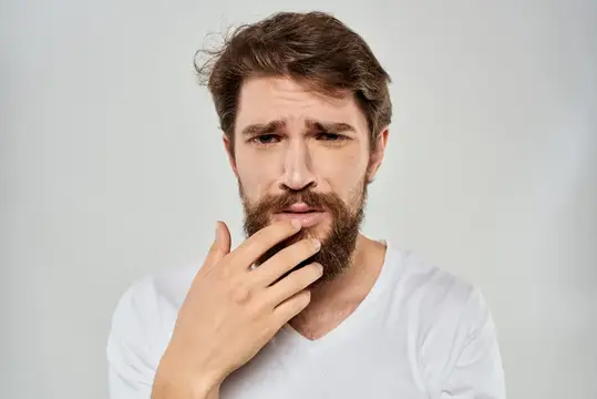 Задумчивый бородатый мужчина прикрывает рот рукой на светлом фоне