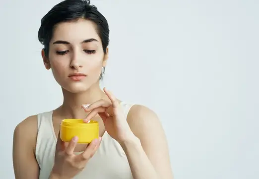 Молодая брюнетка наносит крем из желтой баночки на светлом фоне