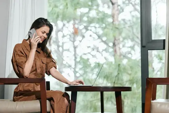 Улыбающаяся брюнетка говорит по телефону за ноутбуком у окна