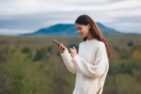 Улыбающаяся девушка в белом свитере смотрит в телефон на фоне гор