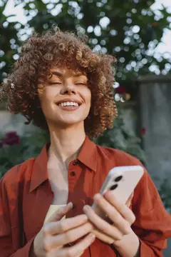 Улыбающаяся женщина на улице пользуется смартфоном, одетая в повседневную одежду в солнечный день