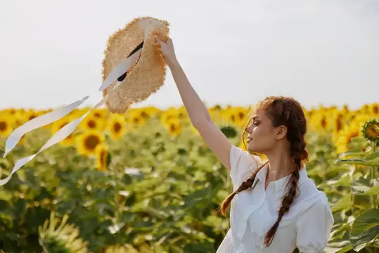 Молодая женщина с косичками поднимает соломенную шляпу в поле подсолнухов