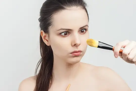 Молодая женщина с кистью для макияжа у щеки на сером фоне