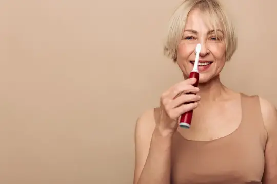 Улыбающаяся седовласая женщина чистит зубы электрической щеткой