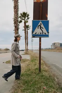 Женщина в плаще и кепке идёт мимо знака пешеходного перехода на фоне пальм