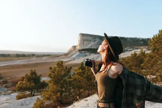 Женщина-фотограф в клетчатой рубашке и шляпе снимает живописный вид с вершины холма