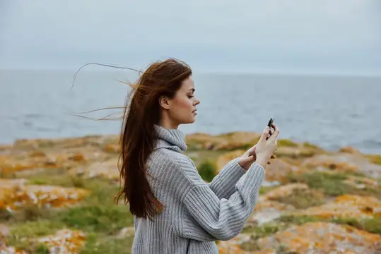Девушка в сером свитере фотографирует море на телефон