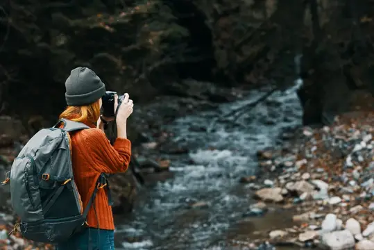 Рыжеволосая девушка с рюкзаком фотографирует горную реку