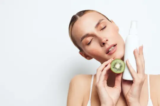 Молодая женщина с закрытыми глазами держит киви и флакон косметики