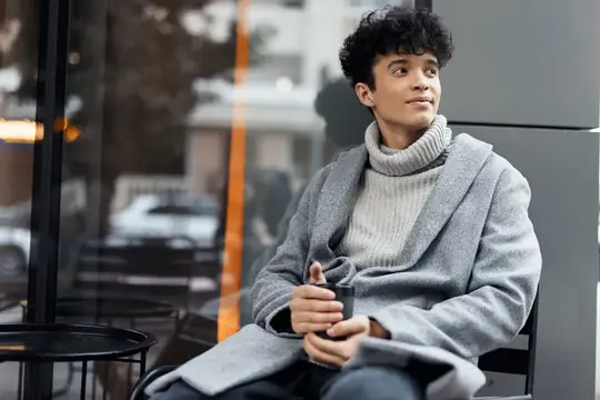 Кудрявый парень в сером пальто с кофе у окна кафе