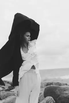Мрачный черно-белый fashion-кадр: женщина, развевающаяся ткань и развевающийся на ветру плащ на фоне сурового побережья, смелая поза и кинематографичное освещение