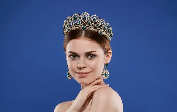 Модель в образе принцессы с короной на голове, макияж, жест рукой, синий фон