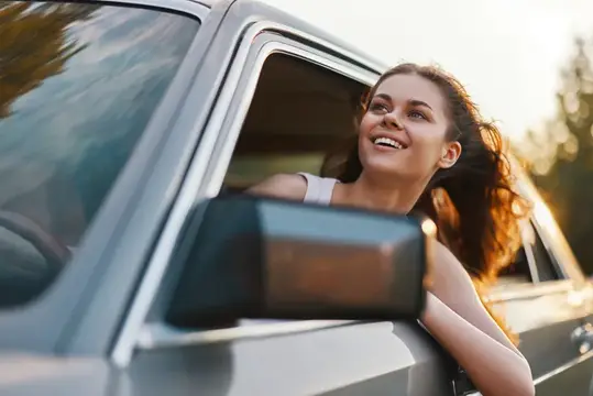 Улыбающаяся девушка высунулась из окна автомобиля на закате