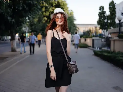 Рыжеволосая девушка в шляпе и солнцезащитных очках гуляет по летней улице