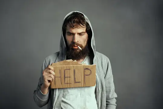 Бородатый мужчина в капюшоне с сигаретой держит табличку Help
