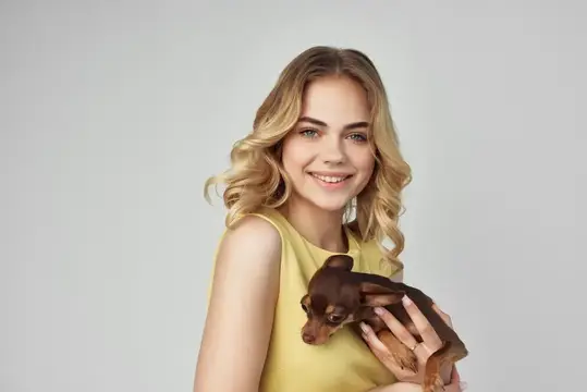 Улыбающаяся блондинка держит маленькую коричневую собачку на сером фоне