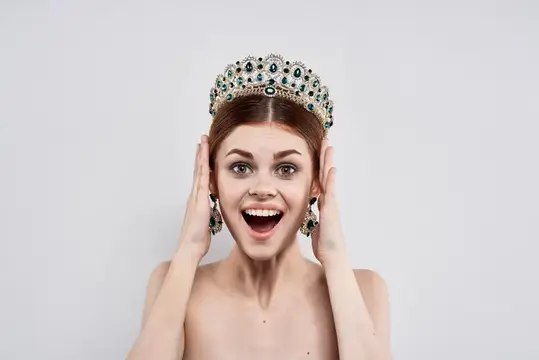 Рыжеволосая девушка в роскошной короне радостно улыбается на сером фоне