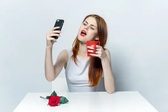 Рыжеволосая девушка с телефоном и красной кружкой за белым столом с розой
