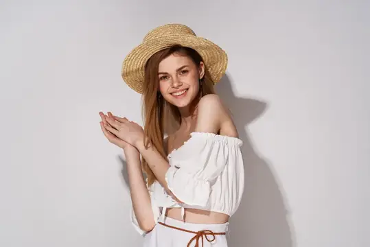 Улыбающаяся девушка в соломенной шляпе и белой блузе на сером фоне