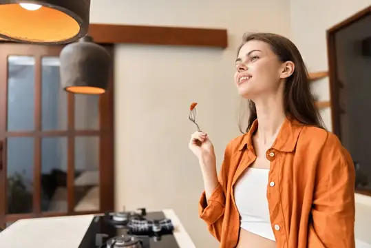 Веселая молодая женщина в оранжевой куртке наслаждается здоровой едой на современной кухне, вызывая чувства тепла и счастья