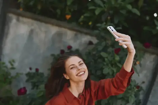 Улыбающаяся темноволосая девушка делает селфи на фоне розовых кустов