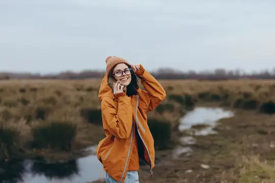 Женщина на открытом воздухе, поле, река, пальто, шляпа, улыбка, очки, зимний образ жизни, портрет у воды на лугу, оранжевое пальто и вязаная шапка-бини, очки видны, жизнерадостное расслабленное…