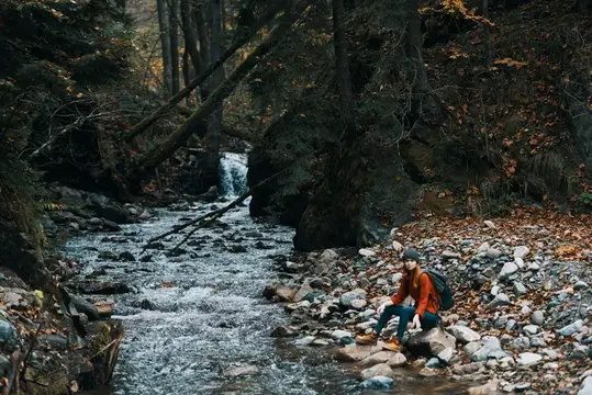 Женщина с рюкзаком на фоне осеннего горного пейзажа, леса и чистой воды в реке