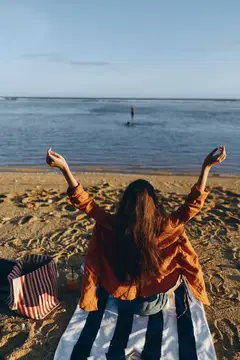 Пляж, океан, женщина с поднятыми руками в золотом свете заката, горизонт с ясным небом, мягкие волны и фактурный песок, длинные вьющиеся волосы, джинсовая куртка, полосатая хозяйственная сумка, на…
