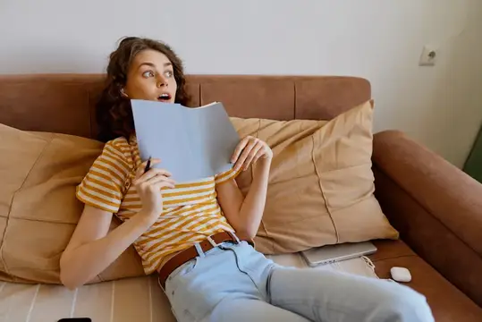 Женщина на диване дома читает книгу с удивлённым выражением лица, в полосатой футболке и джинсах, тёплый спокойный интерьер, дневной свет, расслабленная сцена образа жизни, сосредоточенная на чтении
