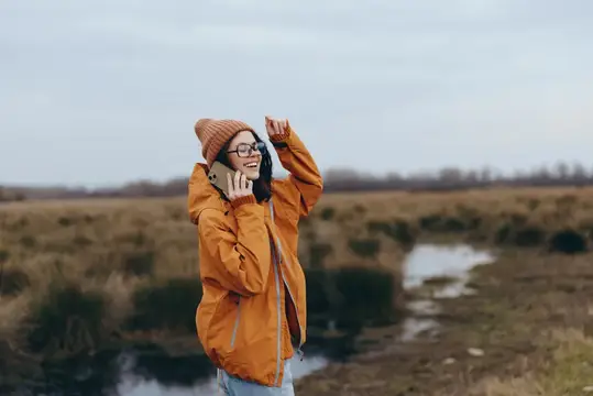 Улыбающаяся девушка в оранжевой куртке и шапке разговаривает по телефону в поле