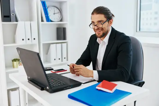 Улыбающийся бородатый мужчина в очках работает за ноутбуком в офисе