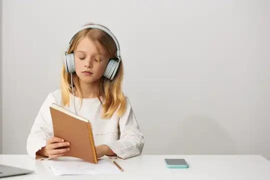 Светловолосая девочка в наушниках читает книгу за столом