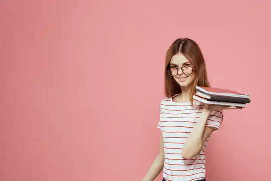 Молодая студентка с книгами на розовом фоне, в очках, крупный план лица