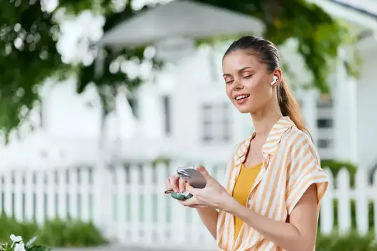 Улыбающаяся девушка в наушниках со смартфоном у белого забора