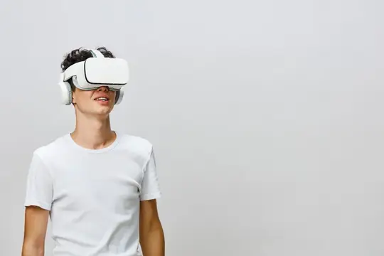 Молодой человек в белой футболке и VR-очках улыбается на сером фоне