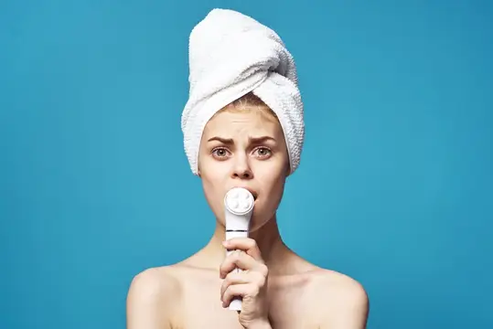 Молодая женщина с полотенцем на голове чистит лицо щеткой на синем фоне