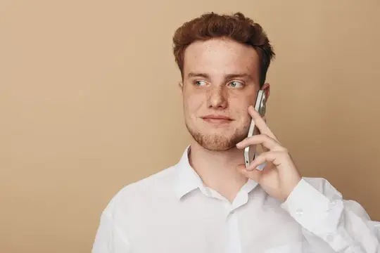 Рыжеволосый мужчина с бородой в белой рубашке держит смартфон у уха и смотрит в сторону на бежевом фоне