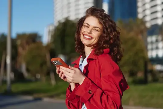 Улыбающаяся кудрявая девушка в красном плаще с телефоном на городской улице