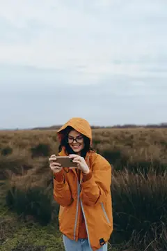 Улыбающаяся девушка в оранжевом дождевике фотографирует на телефон в поле