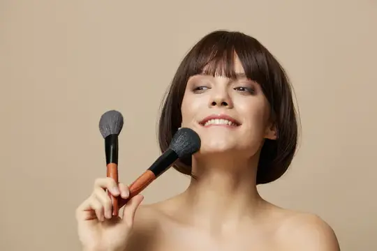 Улыбающаяся девушка с каре держит две кисти для макияжа