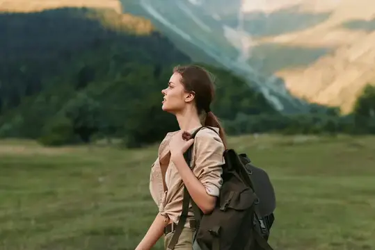 Молодая женщина в бежевой рубашке с большим рюкзаком стоит на зеленой траве на фоне гор, что навевает мысли об исследовании, свободе и безмятежности на природе