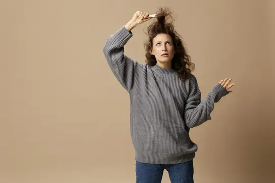 Кудрявая женщина в сером свитере причёсывается на бежевом фоне